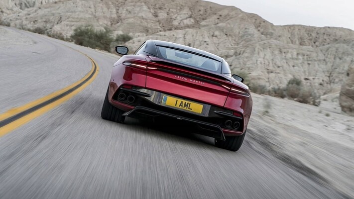 Vista trasera dinámica del DBS Superleggera en carretera, destacando su alerón y luces traseras. Vista trasera dinámica del DBS Superleggera en carretera, destacando su alerón y luces traseras.