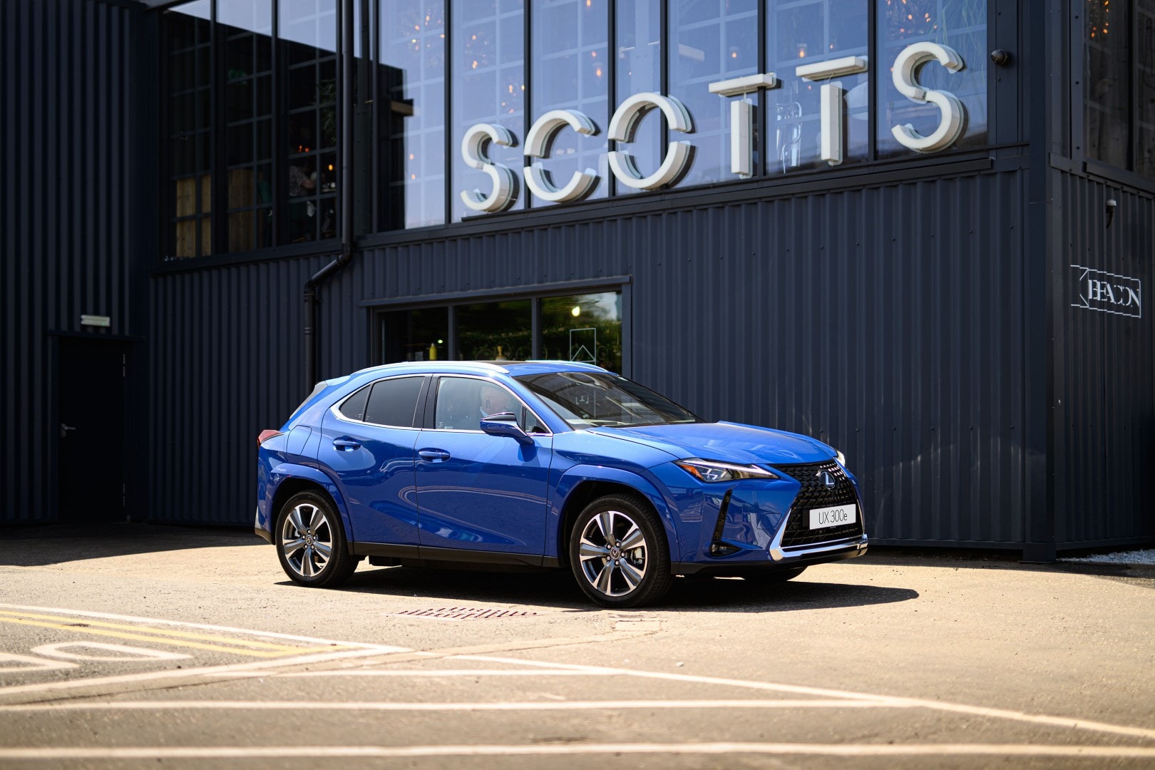 Un Lexus UX300e azul estacionado lateralmente frente a un ambiente moderno.