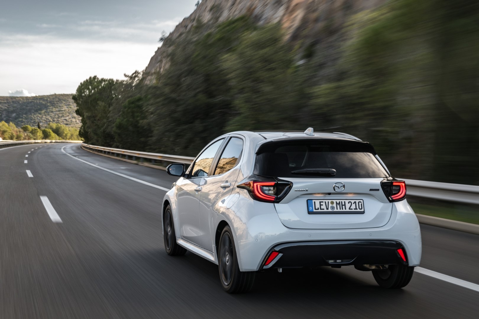 Vista trasera del Mazda 2 híbrido en carretera, destacando su silueta y luces traseras.