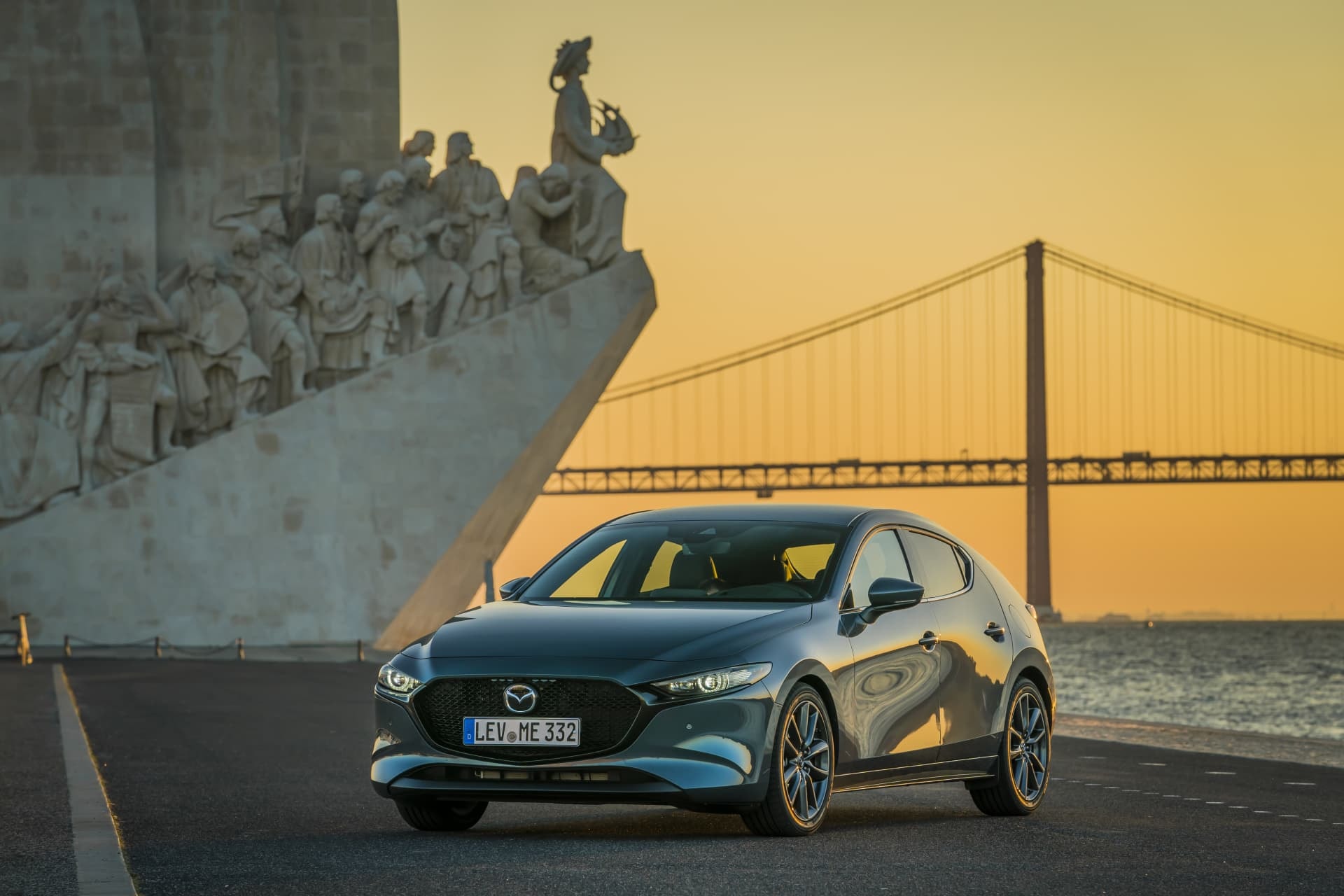 Frente del Mazda 3 en tono gris iluminado por el atardecer, junto a un monumento histórico.