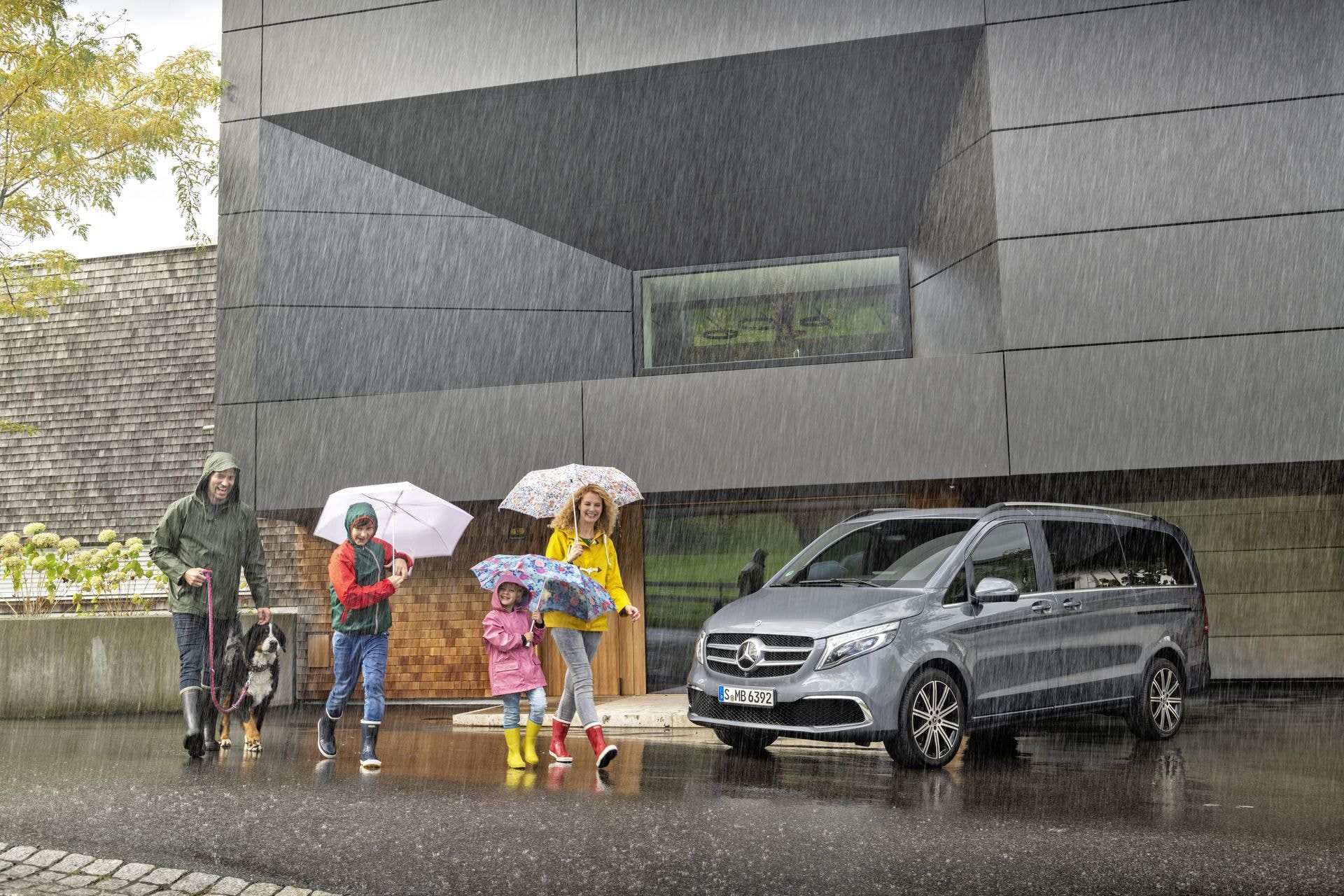 Mercedes Clase V aparcado en día lluvioso, familia con paraguas al lado