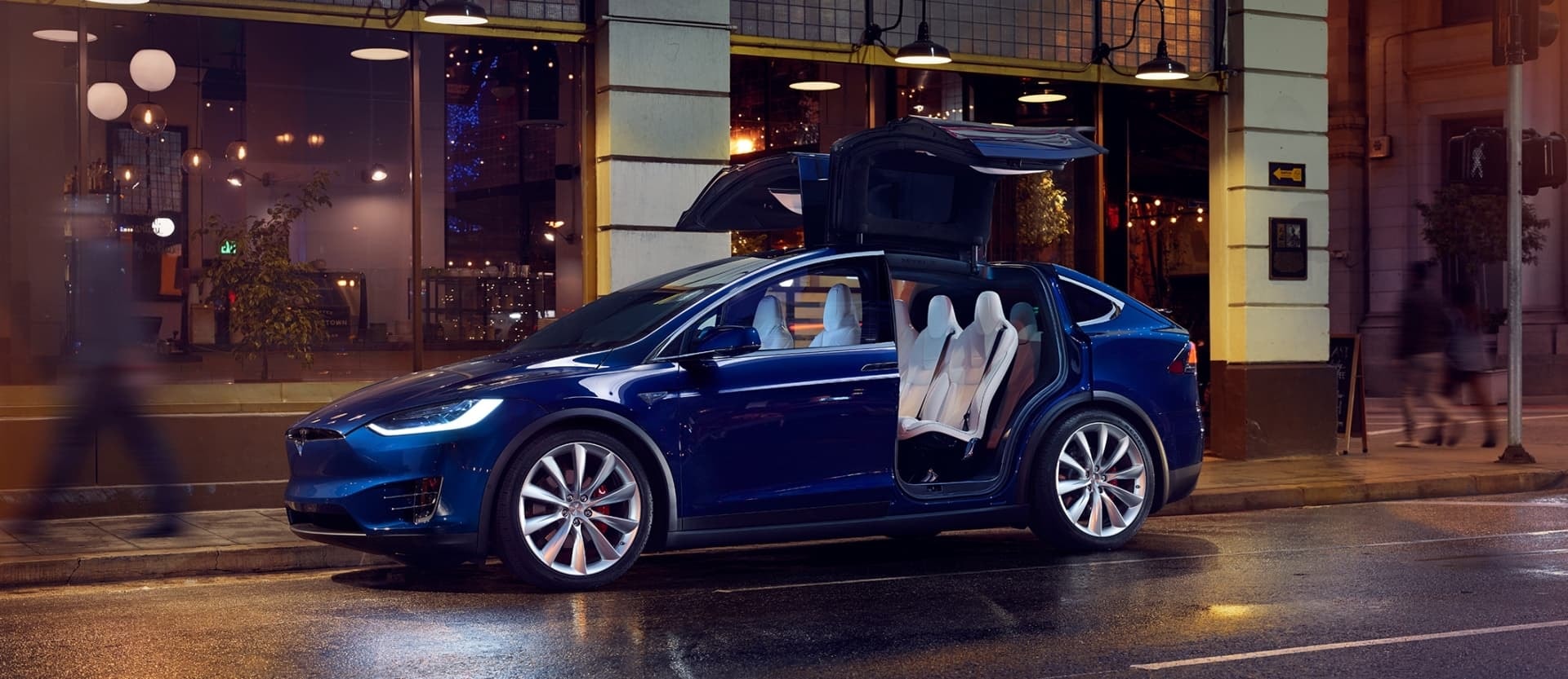 Perfil del Tesla Model X con las puertas de ala de gaviota abiertas en ambiente urbano.